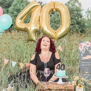 Puede incluir: Una mujer con un vestido negro se sienta en un campo de hierba con un gran globo de oro n&uacute;mero 40 detr&aacute;s de ella. Ella sonr&iacute;e y sostiene una copa de vino. Hay un peque&ntilde;o pastel con un n&uacute;mero 40 en frente de ella. Un letrero de pizarra con el nombre Monica y las palabras "Es mi cumplea&ntilde;os n&uacute;mero 40" est&aacute; en el fondo.