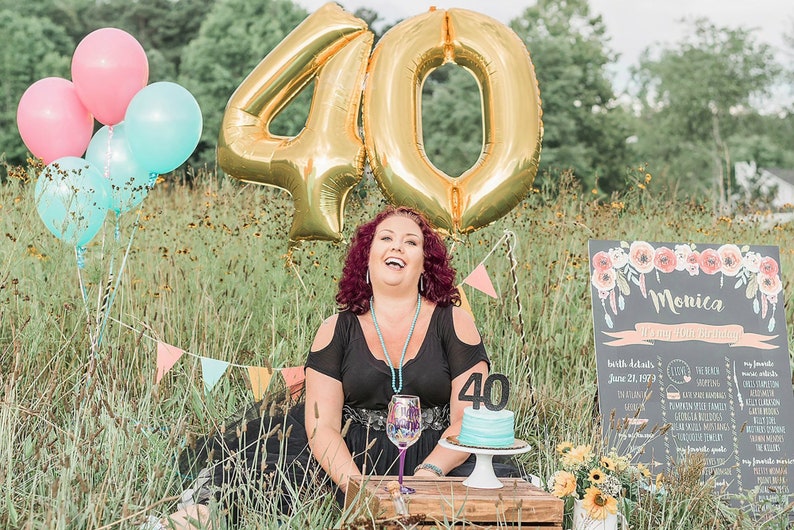 Puede incluir: Una mujer con un vestido negro se sienta en un campo de hierba con un gran globo de oro n&uacute;mero 40 detr&aacute;s de ella. Ella sonr&iacute;e y sostiene una copa de vino. Hay un peque&ntilde;o pastel con un n&uacute;mero 40 en frente de ella. Un letrero de pizarra con el nombre Monica y las palabras "Es mi cumplea&ntilde;os n&uacute;mero 40" est&aacute; en el fondo.