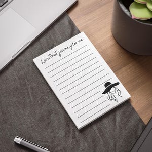 以下が含まれることがあります： 「Love that journey for me」の黒い筆記体で書かれた白い罫線入りのメモ帳。帽子をかぶった女性の白黒イラストが右下にあります。 メモ帳は濃い灰色の表面にあります。
