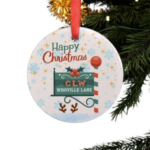 Puede incluir: Un adorno navideño redondo con una cinta roja para colgar. El adorno presenta las palabras "Happy Christmas" en rojo y un letrero verde que dice "CLW Whoville Lane". Copos de nieve y estrellas decoran el fondo.