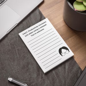 May include: White notepad with lined pages and the text "FACT: Productivity increases 11% when using a notepad. You're welcome." A black and white cartoon image of a person with glasses is in the bottom right corner. The notepad is on a wooden desk.