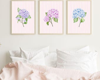 May include: Three framed floral prints featuring watercolor hydrangeas in shades of purple, pink, and blue. Each print has a pink and white striped background. The prints are displayed above a bed with white pillows and a pink blanket.