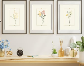 Op de afbeelding: Drie ingelijste botanische kunstprints. Elke print toont een delicaat bloemstuk in een rechthoekige gouden lijst op een gebroken witte achtergrond. De lijsten zijn donkerbruin en het kunstwerk wordt op een witte muur getoond.