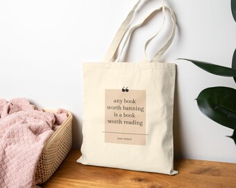 May include: Cream-colored tote bag with the quote "any book worth banning is a book worth reading" in a tan rectangle. The bag has long handles and is sitting on a wooden surface. A basket with a pink blanket is in the background.