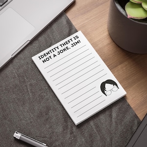 May include: A white lined notepad with the text "IDENTITY THEFT IS NOT A JOKE, JIM!" in black. A black and white cartoon image of a person with glasses is in the bottom right corner. The notepad is on a dark grey surface.
