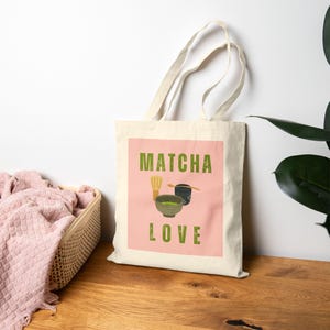 May include: A cream-colored tote bag with a pink rectangle featuring the words "Matcha Love" in green. The design includes illustrations of matcha tea preparation tools: a whisk, a bowl, and a cup.