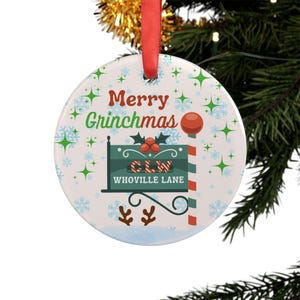 May include: A circular Christmas tree decoration with a red hanging ribbon. The ornament displays the words "Merry Grinchmas" in green and red, and a sign reading "CLW Whoville Lane". The background is decorated with snowflakes and green star accents.