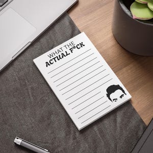 Pode incluir: Bloco de notas branco com linhas pretas e o texto "WHAT THE ACTUAL F*CK". Uma ilustração a preto e branco do rosto de um homem está no canto inferior direito. O bloco de notas está sobre uma superfície cinzenta escura.