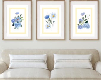 May include: Three framed floral art prints. Each print features a watercolor painting of blue flowers on a white background, set within a yellow border and a wooden frame. The prints are displayed above a beige sofa with striped pillows.
