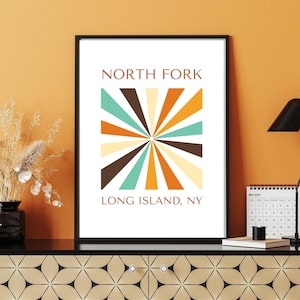 May include: Framed art print featuring the text "NORTH FORK LONG ISLAND, NY." The artwork has a retro design with a burst of colorful rays in brown, teal, orange, and cream. The frame is black, and the background is a warm orange.
