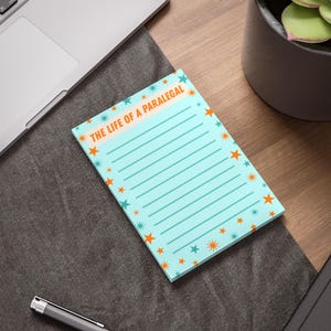 May include: A rectangular notepad with a light blue background featuring a repeating pattern of orange and teal stars. The top section contains the text THE LIFE OF A PARALEGAL in orange capital letters, followed by a series of horizontal lines.