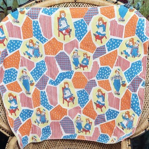 May include: A vintage fabric with a repeating pattern of dolls in blue and white outfits, sitting on chairs. The background features orange, red, and blue geometric shapes. The fabric is draped over a wicker chair.