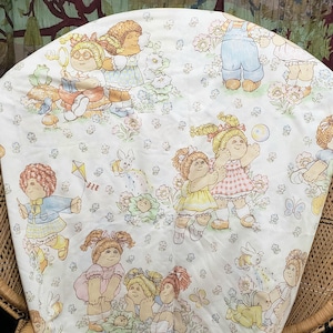 May include: A white fabric featuring a repeating pattern of Cabbage Patch Kids dolls in various outfits, including overalls, dresses, and sweaters. The illustrations are rendered in soft pastel colors, depicting the dolls playing with kites, bubbles, and flowers.