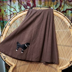 May include: A brown wool skirt with a black poodle applique. The skirt is a-line and has a high waist. It is sitting on a wicker chair.