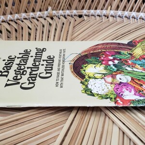 May include: A gardening guide titled "Basic Vegetable Gardening Guide" with a colorful illustration of various vegetables. The book is lying on a woven, light brown surface.