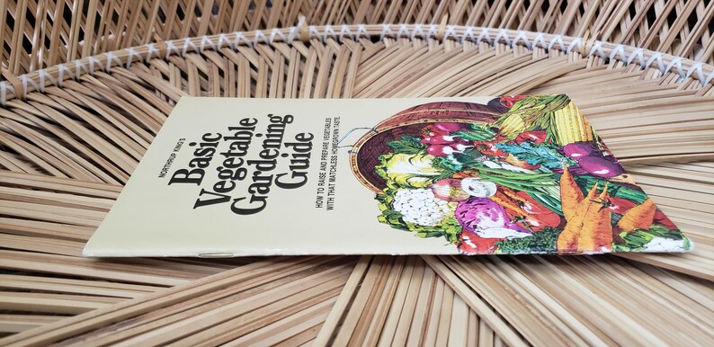 May include: A gardening guide titled "Basic Vegetable Gardening Guide" with a colorful illustration of various vegetables. The book is lying on a woven, light brown surface.