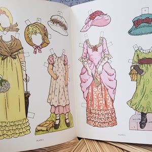 Puede incluir: Libro abierto con mu&ntilde;ecas de papel vintage. Las mu&ntilde;ecas visten vestidos y gorros hist&oacute;ricos, con una variedad de colores como amarillo, rosa y verde. Las ilustraciones son detalladas y el libro est&aacute; abierto en una doble p&aacute;gina.
