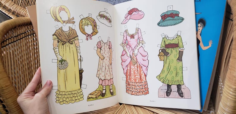 Puede incluir: Libro abierto con mu&ntilde;ecas de papel vintage. Las mu&ntilde;ecas visten vestidos y gorros hist&oacute;ricos, con una variedad de colores como amarillo, rosa y verde. Las ilustraciones son detalladas y el libro est&aacute; abierto en una doble p&aacute;gina.