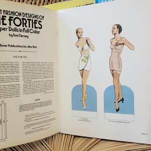 Puede incluir: Libro abierto titulado "Great Fashion Designs of the Forties" con ilustraciones de mu&ntilde;ecas de papel. El libro presenta dibujos de mujeres con ropa vintage. A la derecha se ve un libro azul con una mujer con un vestido negro.