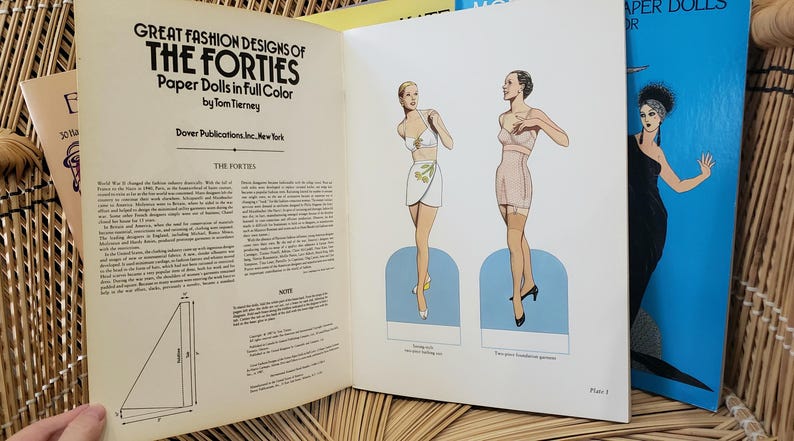 Puede incluir: Libro abierto titulado "Great Fashion Designs of the Forties" con ilustraciones de mu&ntilde;ecas de papel. El libro presenta dibujos de mujeres con ropa vintage. A la derecha se ve un libro azul con una mujer con un vestido negro.