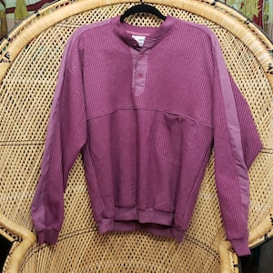 May include: A mauve-colored long-sleeved shirt with a ribbed texture, a button-up collar, and a small pocket. The shirt is hanging on a black hanger against a wicker chair.