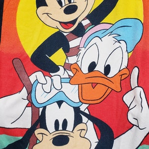 May include: A colourful beach towel featuring Mickey Mouse, Donald Duck, and Goofy. Mickey is wearing sunglasses and a striped shirt. Donald is wearing a blue hat and a white shirt. Goofy is wearing goggles and a pink shirt. The towel has the text "St. Thomas Virgin Islands" at the top and "Disney" at the bottom.