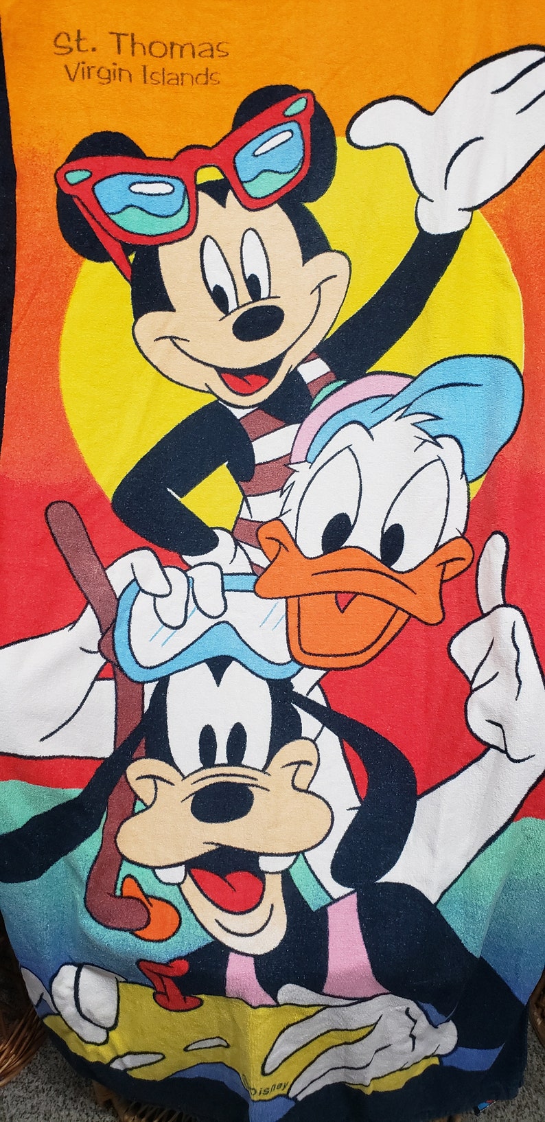 May include: A colourful beach towel featuring Mickey Mouse, Donald Duck, and Goofy. Mickey is wearing sunglasses and a striped shirt. Donald is wearing a blue hat and a white shirt. Goofy is wearing goggles and a pink shirt. The towel has the text "St. Thomas Virgin Islands" at the top and "Disney" at the bottom.