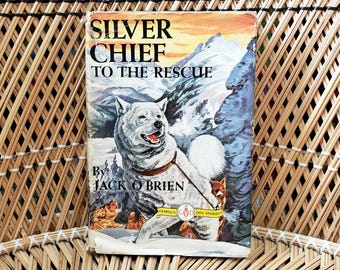 May include: A vintage book titled "Silver Chief to the Rescue" by Jack O'Brien. The cover features a white dog with a mountain backdrop. The book is set against a woven background.