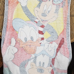 May include: A white beach towel featuring a colourful cartoon image of Mickey Mouse, Donald Duck and Goofy. Mickey Mouse is wearing red sunglasses and a red and white striped shirt. Donald Duck is wearing a blue and white hat. Goofy is wearing a blue and white hat and a blue and white shirt.