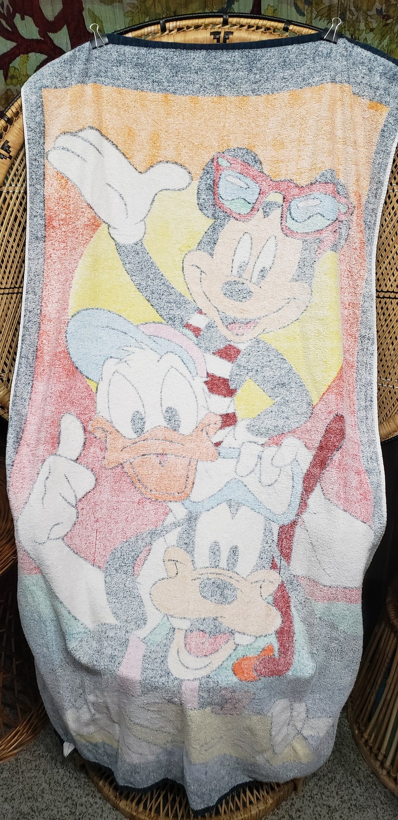 May include: A white beach towel featuring a colourful cartoon image of Mickey Mouse, Donald Duck and Goofy. Mickey Mouse is wearing red sunglasses and a red and white striped shirt. Donald Duck is wearing a blue and white hat. Goofy is wearing a blue and white hat and a blue and white shirt.