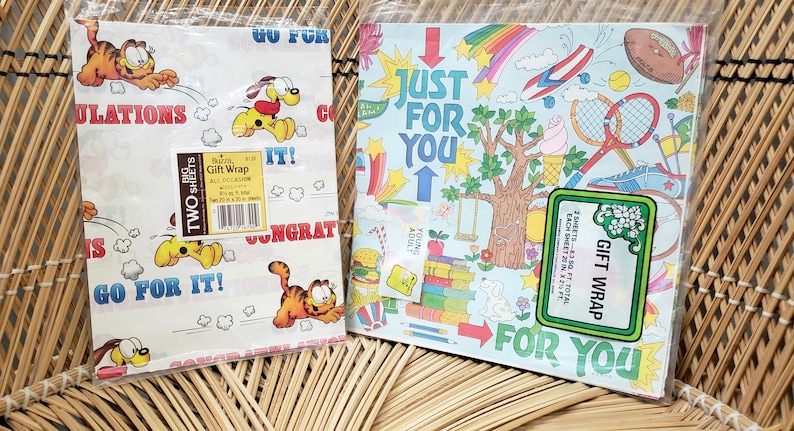 Puede incluir: Dos hojas de papel de regalo con el tema de Garfield. Una hoja presenta a Garfield y Odie con el texto "Go For It!" y "Congratulations". La otra hoja presenta un arco&iacute;ris, nubes y el texto "Just For You".