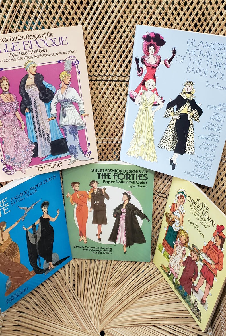 Puede incluir: Una colecci&oacute;n de libros de mu&ntilde;ecas de papel vintage con dise&ntilde;os de moda de varias &eacute;pocas. Las portadas muestran ilustraciones de figuras elegantes con atuendos coloridos, con t&iacute;tulos como "Great Fashion Designs" y "Glamorous Movie Stars".