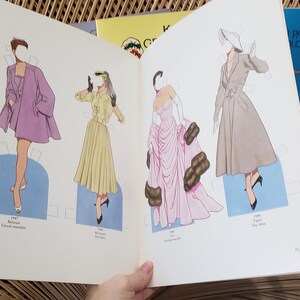 Puede incluir: Un libro abierto de mu&ntilde;ecas de papel vintage con ilustraciones de moda. Las mu&ntilde;ecas visten un abrigo morado, un vestido amarillo y un vestido rosa. Se ve un libro azul titulado "Paper Dolls".