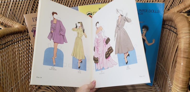 Puede incluir: Un libro abierto de mu&ntilde;ecas de papel vintage con ilustraciones de moda. Las mu&ntilde;ecas visten un abrigo morado, un vestido amarillo y un vestido rosa. Se ve un libro azul titulado "Paper Dolls".
