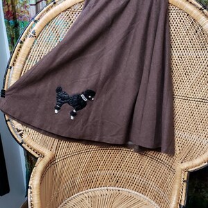May include: A brown skirt with a black poodle applique. The skirt is hanging on a wicker chair.