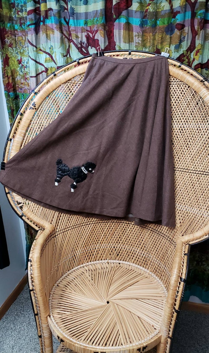 May include: A brown skirt with a black poodle applique. The skirt is hanging on a wicker chair.
