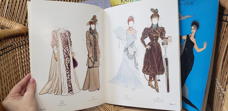 Puede incluir: Libro abierto con mu&ntilde;ecas de papel vintage. Las mu&ntilde;ecas visten trajes hist&oacute;ricos, incluyendo vestidos largos, abrigos y sombreros. Las ilustraciones son a color y presentan dise&ntilde;os detallados. Se ve un libro azul con una mujer vestida de negro.