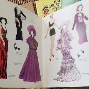 Puede incluir: Libro abierto de mu&ntilde;ecas de papel vintage con ilustraciones de moda. Las p&aacute;ginas muestran dibujos de mujeres con varios vestidos, incluyendo un vestido rojo con lentejuelas y un vestido morado. Otros libros son visibles en el fondo.