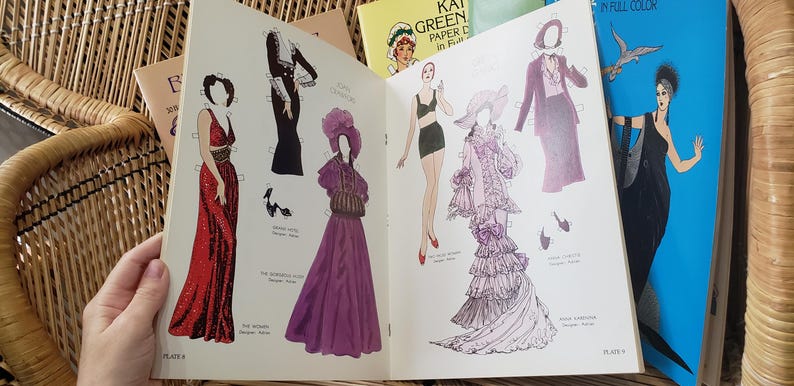 Puede incluir: Libro abierto de mu&ntilde;ecas de papel vintage con ilustraciones de moda. Las p&aacute;ginas muestran dibujos de mujeres con varios vestidos, incluyendo un vestido rojo con lentejuelas y un vestido morado. Otros libros son visibles en el fondo.