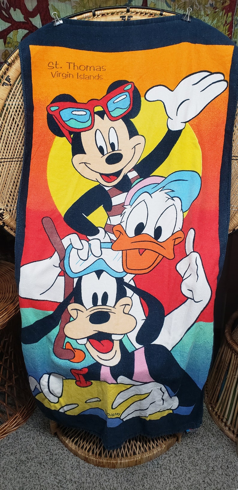May include: A colourful beach towel featuring Mickey Mouse, Donald Duck, and Goofy wearing sunglasses and enjoying the beach. The towel has the text "St. Thomas Virgin Islands" at the top.
