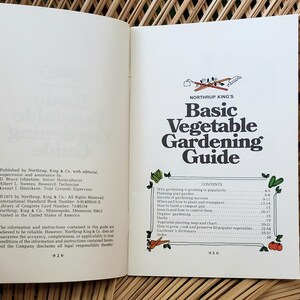 May include: Open book with the title "Basic Vegetable Gardening Guide" on the right page. The left page has text about the publisher. The book is open on a woven surface.
