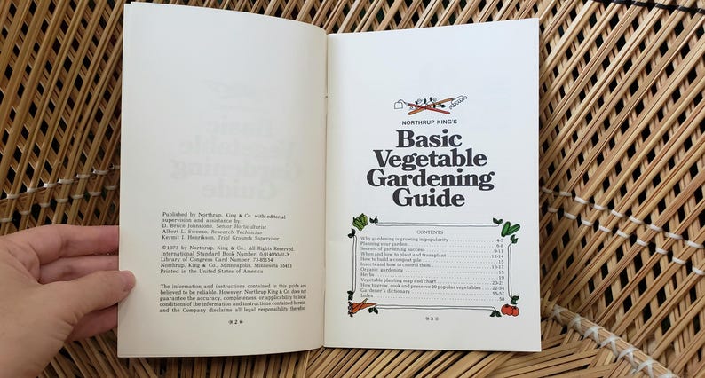 May include: Open book with the title "Basic Vegetable Gardening Guide" on the right page. The left page has text about the publisher. The book is open on a woven surface.