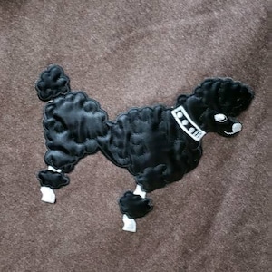 May include: A brown felt throw blanket with a black embroidered poodle design. The poodle has a white collar with black dots.