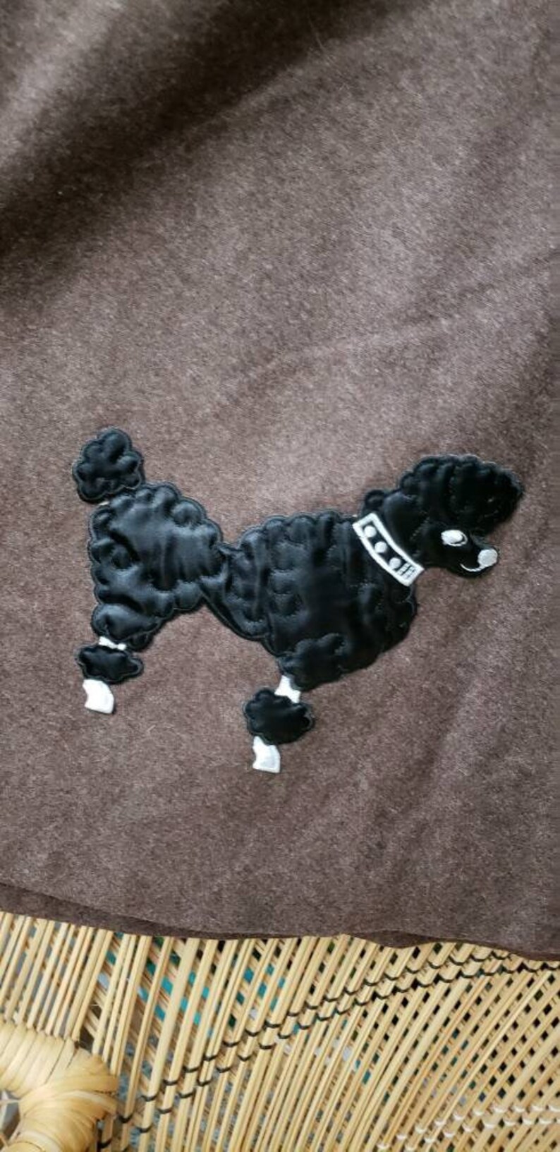 May include: A brown felt throw blanket with a black embroidered poodle design. The poodle has a white collar with black dots.