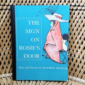 May include: A blue book with the title "The Sign on Rosie's Door" by Maurice Sendak. The cover features a girl wearing a red cloak and a white hat with a feather, sitting on a chair with a white cat.