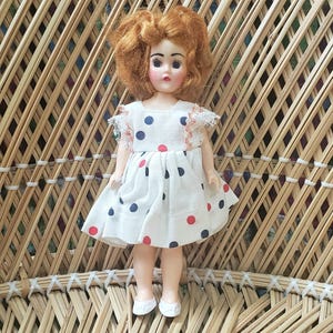 May include: A vintage doll with red hair and a white dress adorned with red and blue polka dots. The doll has a sweet expression and is wearing white shoes. The doll is sitting on a woven wicker chair.