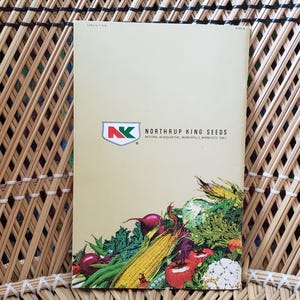 May include: A seed catalog with the text "Northrup King Seeds" and a logo featuring a red and green design. The catalog displays a colorful illustration of various vegetables, including corn, tomatoes, and cauliflower. The catalog is set against a woven background.