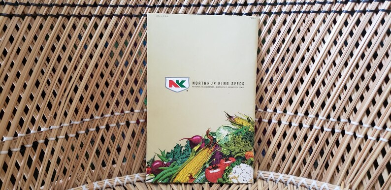 May include: A seed catalog with the text "Northrup King Seeds" and a logo featuring a red and green design. The catalog displays a colorful illustration of various vegetables, including corn, tomatoes, and cauliflower. The catalog is set against a woven background.