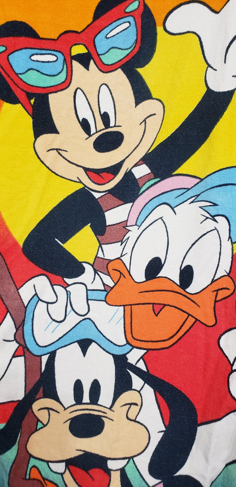 May include: A colourful illustration of Mickey Mouse, Donald Duck, and Goofy wearing sunglasses and swimming gear. Mickey is wearing red sunglasses, Donald Duck is wearing a blue and white swimming cap, and Goofy is wearing blue goggles. The background is a yellow and orange ombre.