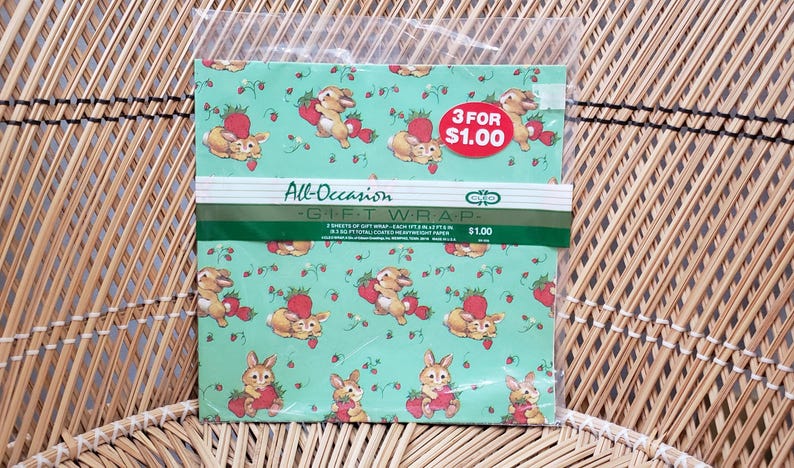 Puede incluir: Una hoja de papel de regalo verde con un patr&oacute;n de conejos y fresas. El texto "All-Occasion Gift Wrap" y "3 por $1.00" est&aacute; impreso en la hoja.